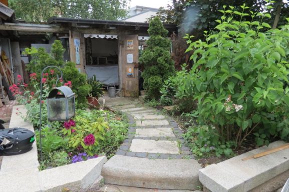 Ein Gartenweg mit Blumenbeeten und einem kleinen Schuppen im Hintergrund.