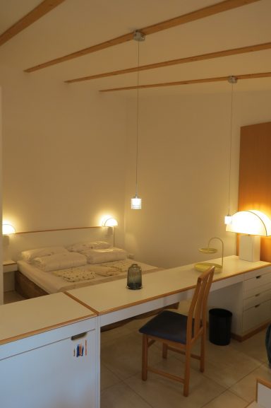 Helles Zimmer mit Doppelbett, Schreibtisch, Stuhl und Nachttischlampen.